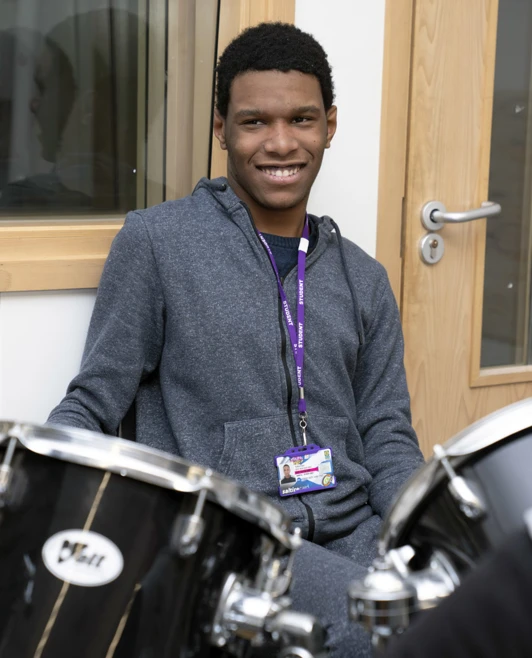 Young man sitting and smiling next to a drum kit in a music room, wearing a lanyard. Young man sitting and smiling next to a drum kit in a music room, wearing a lanyard.